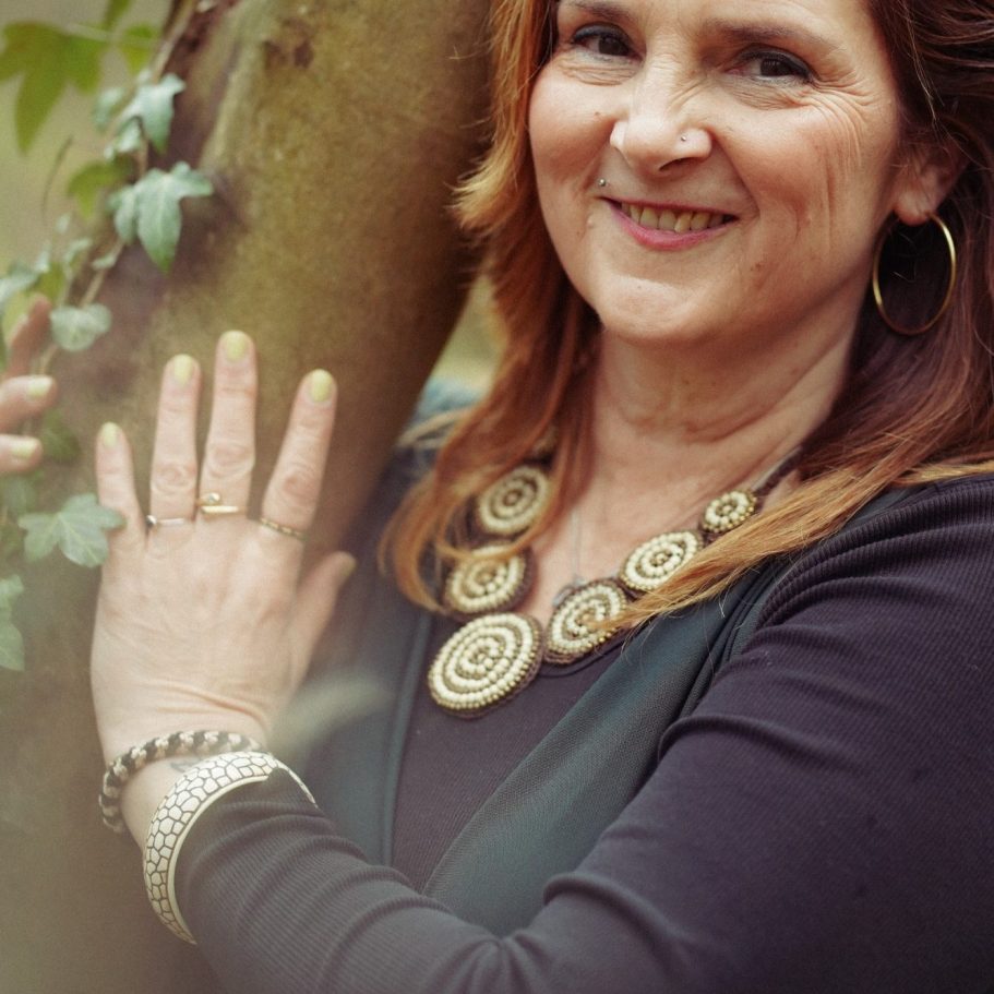 Frau mit langen roten Haaren, die an einem Baum steht und lächelt.